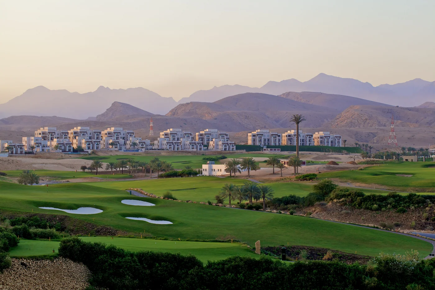 Villas blancas a lo lejos con vistas a jardines verdes y montañas detrás de Muscat Hills, Omán.